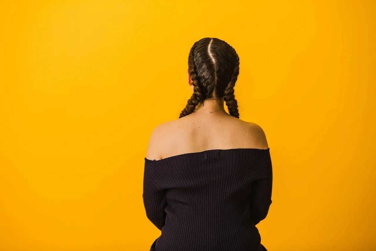 Person with braided hair facing a bright yellow wall, wearing an off-shoulder black top.