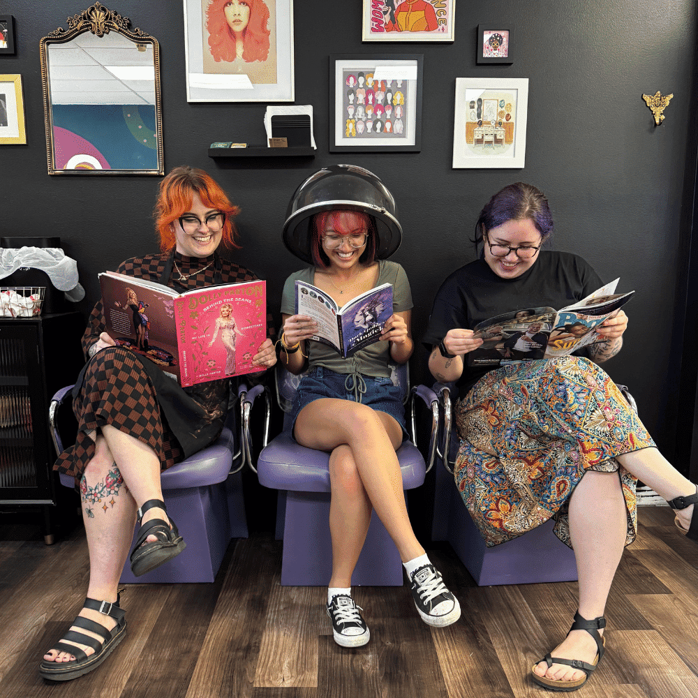 Three people with colorful hairstyles read magazines in a salon waiting area, smiling.